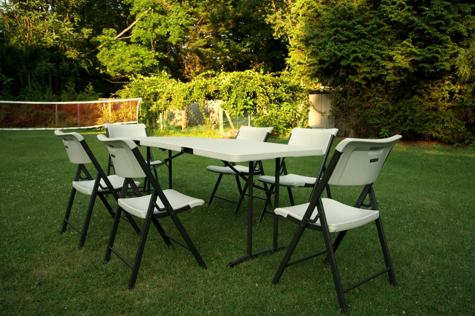 Six-foot folding table with six white folding chairs set up on a Connecticut backyard lawn at golden hour